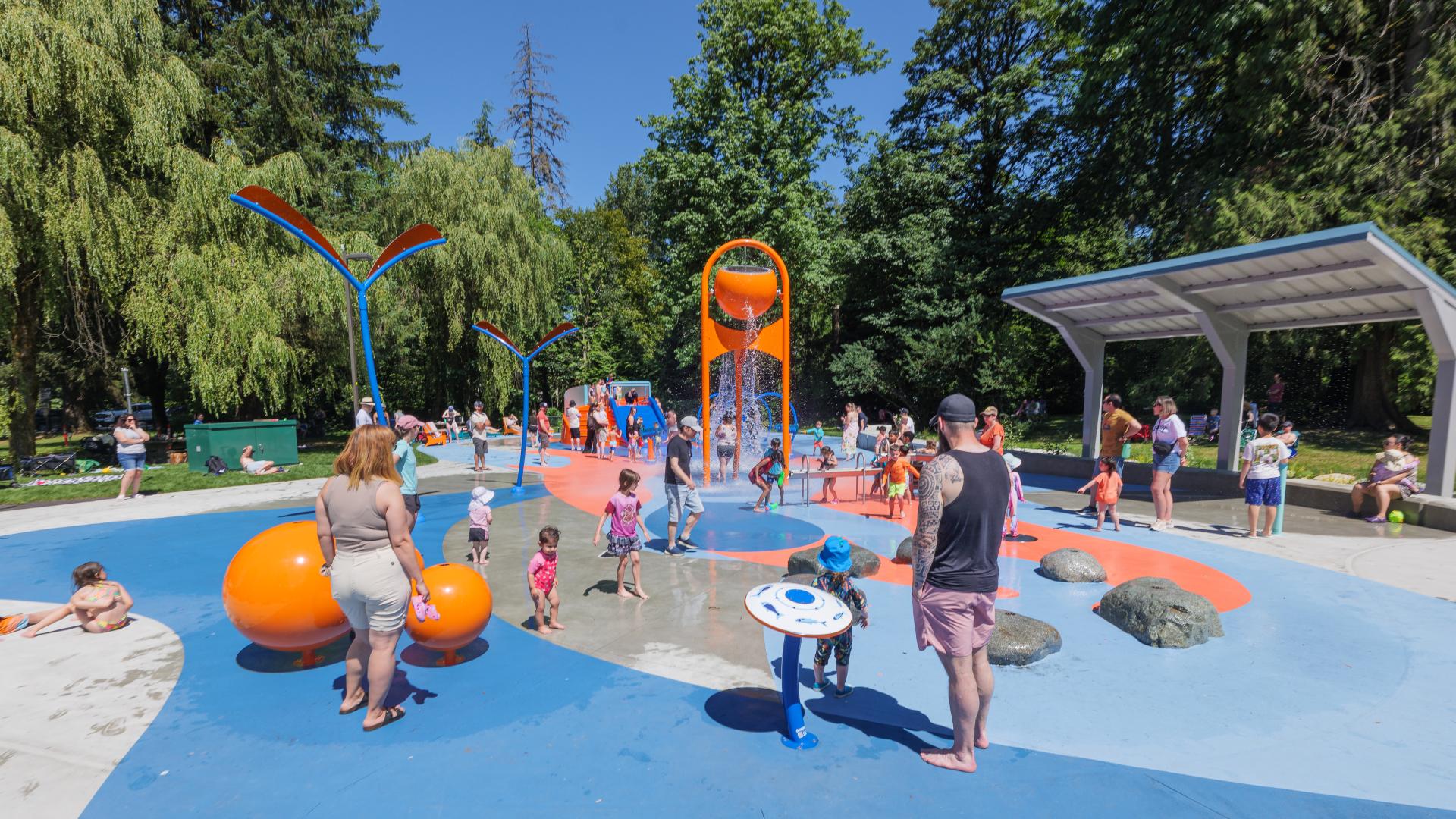 A spray park with blue painted concrete deck, orange spray features and a metal water dripping shelter
